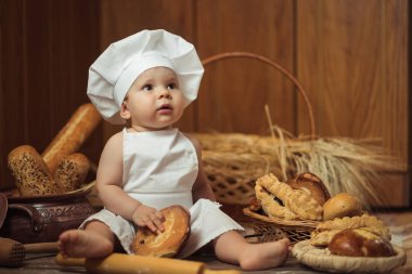küçük çocuk Kostüm giymiş bir aşçı un şekerlemeler kahverengi ahşap zemin üzerinde bir fırın ekmek rulo arasında bir ahşap arka plan üzerinde oturur