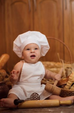 küçük çocuk Kostüm giymiş bir aşçı un şekerlemeler kahverengi ahşap zemin üzerinde bir fırın ekmek rulo arasında bir ahşap arka plan üzerinde oturur