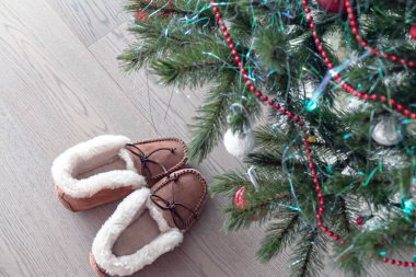 Ev yapımı sıcak kürk terlikler süslenmiş bir Noel ağacının altında yerde duruyor.