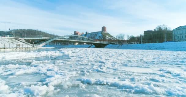 AERIAL. Гладкий полет над рекой Нерис, покрытый льдом в холодный зимний день в столице Литвы Вильнюсе. Панорама замкового холма Гедиминас и моста Миндаугс. 4k — стоковое видео