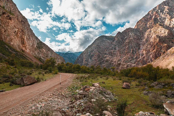 Panorama silnice do rokle hor na pozadí krásných zasněžených vrcholků hor za oblačného počasí. Horská cesta v lese. Pohled na stezku na pozadí modré oblohy — Stock fotografie