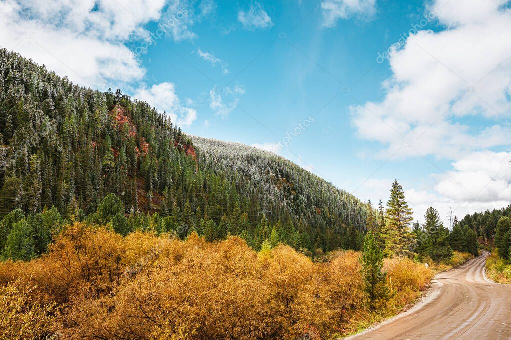 Panorama of road to the gorge of mountains on background of beautiful snowy mountain peaks in cloudy weather. Mountain road in the forest. View of trail on the background of blue sky