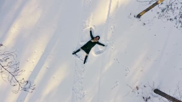 Flicka gör snö ängel i gläntan i skogen. Flygbilder bild av flicka glad att göra med vapen snö ängel figur och ligger i snö. Ung kvinna ligger i gör i skogen — Stockvideo