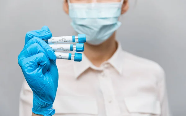 Девушка-врач в медицинской маске и резиновых перчатках, держит в руках, крупным планом пробирки с тестами на наличие ковида-19. Врач в медицинской одежде демонстрирует пробирки с коронавирусом — стоковое фото
