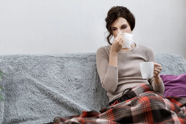 Больной женщины, подозреваемой в ковид-19, находясь дома в постели, измеряет температуру и чихает. Больной женщины, сидящей под одеялом. Девочка на карантине с подозрением на коронавирус — стоковое фото