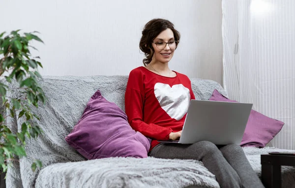 若い女の子は家に隔離され、ソファでリラックスし、コンピュータを使用しています。女はソファに横になってノートパソコンを覗き込む。ガジェットを使用して自宅で休む女の子 — ストック写真