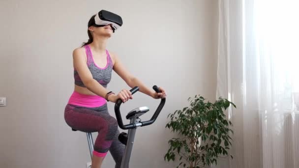 Giovane ragazza rimanere a casa in quarantena e andare in per lo sport con occhiali di realtà virtuale. Donna esercizio yoga mentre a casa indossando occhiali VR. Ragazza rimanere a casa e impegnata in bici stazionaria in VR occhiali — Video Stock