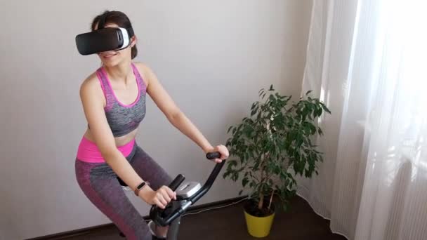 若い女の子は隔離された家に滞在し、仮想現実の眼鏡をかけたスポーツのために行く。VRメガネをかけながら自宅でヨガをする女。女の子は家にいますし、 VRメガネで静止自転車に従事 — ストック動画