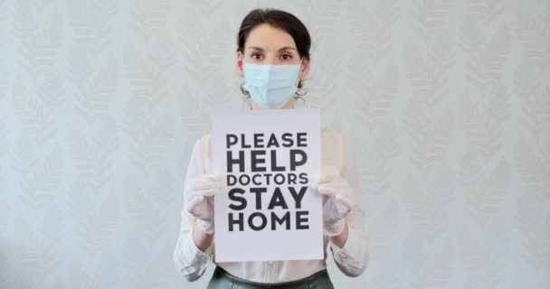 Young girl in medical mask, being at home, to urge help to doctors, showing poster with the inscription, stay home. Woman in gloves showed the inscription, please help doctors, stay at home — Stock Video