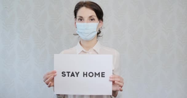 Junges Mädchen in medizinischer Maske, zu Hause, um Ärzte um Hilfe zu bitten, zeigt ein Plakat mit der Aufschrift "Bleibt zu Hause". Frau in Handschuhen zeigte Aufschrift: Bitte helfen Sie Ärzten, bleiben Sie zu Hause — Stockvideo