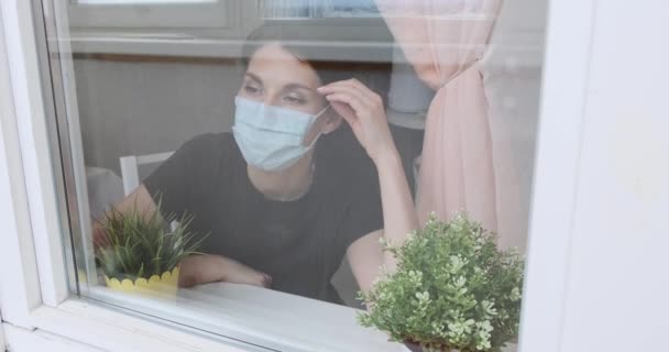 Een jong meisje met een medisch masker, thuis, bij het raam, en naar buiten kijkend door het glas. Vrouw bij het raam in quarantaine tijdens pandemie covid-19, toont een inscriptie verblijf thuis — Stockvideo