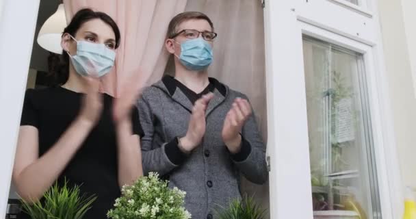 Mladí lidé v lékařských maskovaných potlescích v okně na podporu lidí, kteří bojují proti koronaviru. Muž a žena tleskají z balkónu, na podporu lékařů pomáhajících v boji pandemické covid-19 — Stock video