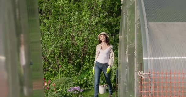 Gyönyörű lány kalapban dolgozik a kertben, belép az üvegházba. A fiatal női farmer ajtót nyit az üvegházba. A dolgozó magánháztartásban dolgozik. A kertész elégedett a munkájával a napsütéses napokon. — Stock videók