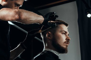 BARBERshop teması. Koyu renk eldivenli sakallı, ciddi bir barber, genç ve yakışıklı müşterisinin saçlarını buduyor. Bir makas ve kostüm kullanıyor.