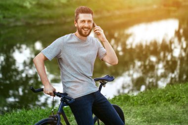 Gri tişörtlü ve mavi kotlu yakışıklı genç adam bisiklet sürmeyi bırakıp, yeşil ağaçlarla dolu bir sokakta, gölün yanında konuşmak istiyor.