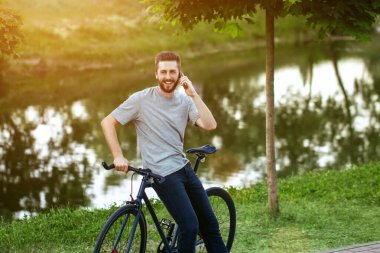 Gri tişörtlü ve mavi kotlu yakışıklı genç adam bisiklet sürmeyi bırakıp, yeşil ağaçlarla dolu bir sokakta, gölün yanında konuşmak istiyor.