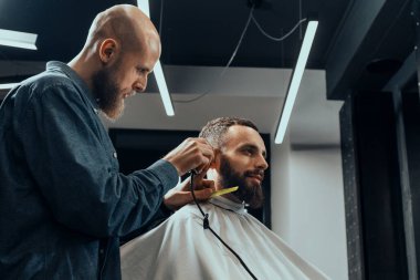 BARBERshop teması. Kel Barber, sakallı, ciddi müşterisinin saçlarını kesiyor. - Kel mi? O bir saç kesicisi kullanıyor.