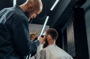 BARBERshop teması. Kel Barber, sakallı, ciddi müşterisinin saçlarını kesiyor. - Kel mi? O bir saç kesicisi kullanıyor.