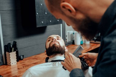 BARBERshop teması. Kel Sakallı BARBER, genç ve yakışıklı müşterisinin sakalını kesiyor. O bir saç kesicisi kullanıyor.