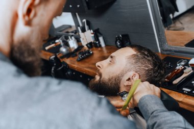 BARBERshop teması. Kel Sakallı BARBER, genç ve yakışıklı müşterisinin sakalını kesiyor. O bir saç kesicisi kullanıyor.