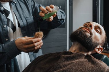 BARBERshop teması. Kel Sakallı Kel, genç ve yakışıklı müşterisi için bir fırçaya tıraş kremi döktü.