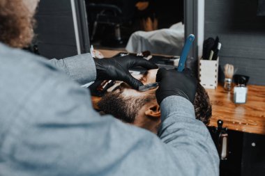 BARBERshop teması. Koyu renk eldivenli BARBER BARBER, genç ve yakışıklı müşterisinin sakalını kesiyor. Heteroseksüel bir RAZOR kullanıyor.