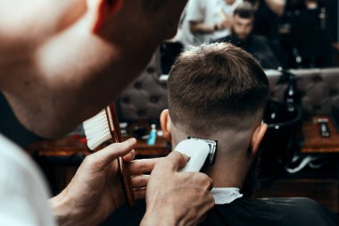 BARBERshop teması. BARBER, GENÇ SİZİ müşterisinin Kıllarını TURTUYOR. O bir saç kesicisi kullanıyor.