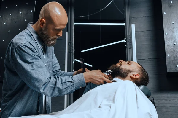 BARBERSHOP THEME. BALD BEARDED BARBER IS TRIMMING THE BEARD OF HIS YOUNG HANDSOME CLIENT. HE IS USING A HAIR CLIPPER