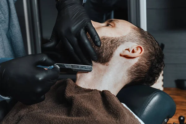 BARBERSHOP THEME. BARBER IN BLACK RUBBER GLOVES IS TRIMMING THE BEARD OF HIS YOUNG HANDSOME CLIENT. HE IS USING A  STRAIGHT RAZOR