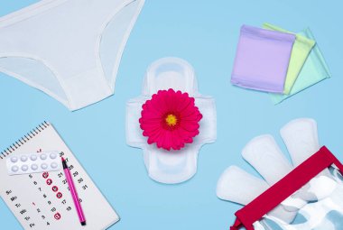 Gerbera çiçeğinin, takvimin, tabletlerin ve regl pedlerinin yanında beyaz bir erkek pantolonu. Kritik günler. Adet görsünler.