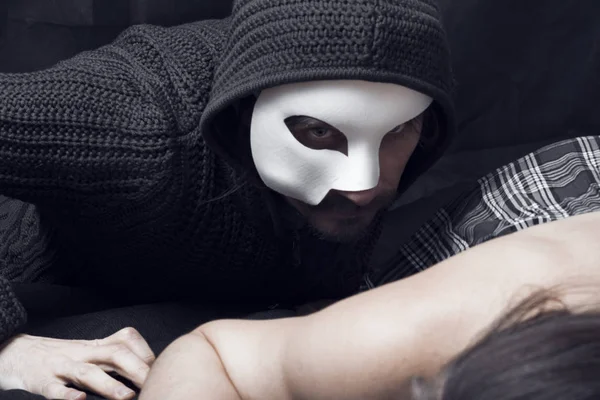 フード付きの黒いセーターを着た白いマスクの男がベッドの上に寝そべっている女の子に這う — ストック写真