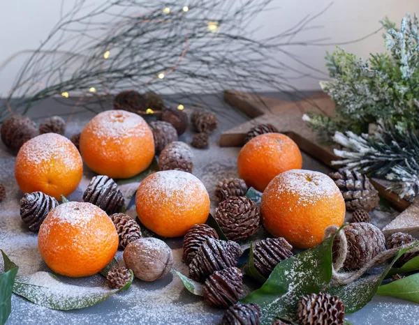 Orangefarbene Mandarinen auf grauem Hintergrund im Neujahrsdekor mit braunen Tannenzapfen und grünen Blättern. Weihnachtsdekoration mit Mandarinen. leckere süße Clementine. — Stockfoto