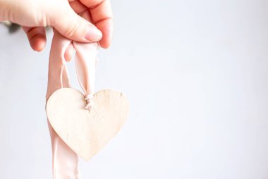 Beyaz arka planda açık ahşap bir kalp ve ipek pembe kurdele. Mesaj için yer var. Soluk pembe bir kurdelenin üzerinde tahta bir kalp kolyesi tutuyor. Boşluğu kopyala.