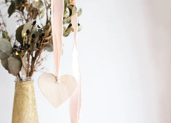 Licht houten hart hanger op een roze satijnen lint op een achtergrond van een gouden glanzende glanzende vaas met gedroogde eucalyptus takken. — Stockfoto