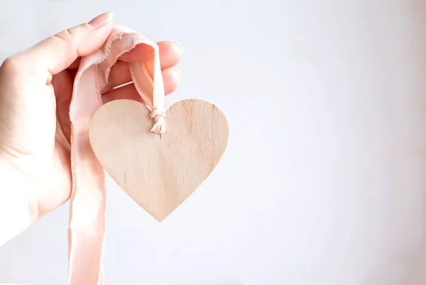 Licht houten hart op een zijden roze lint op witte achtergrond. Ruimte voor tekst. Hand met een houten hart hanger op een chiffon lichtroze lint. Kopieerruimte. — Stockfoto