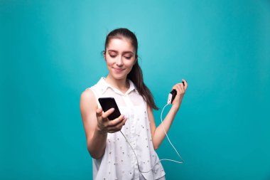 Elinde telefon ve güç bankası olan genç bir kız elinde telefonla gülümsüyor ve elinde mavi cyan arka plan stüdyosu var.