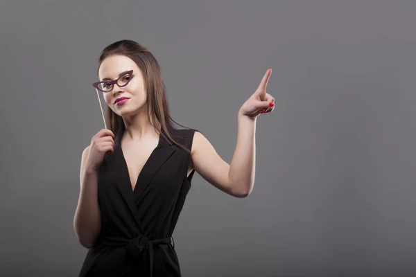 Fille tenant lunettes en papier drôle sur bâton isolé, fond gris. Joyeux jeune femme de la mode. montrant l'espace de copie vide — Photo