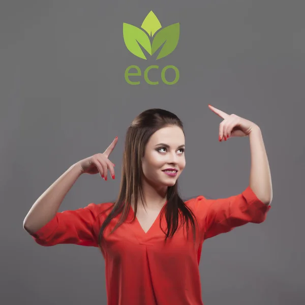 Girl in red shirt, gray background. Joyful young fashion brunette woman. Showing on eco logo — Stock Photo, Image
