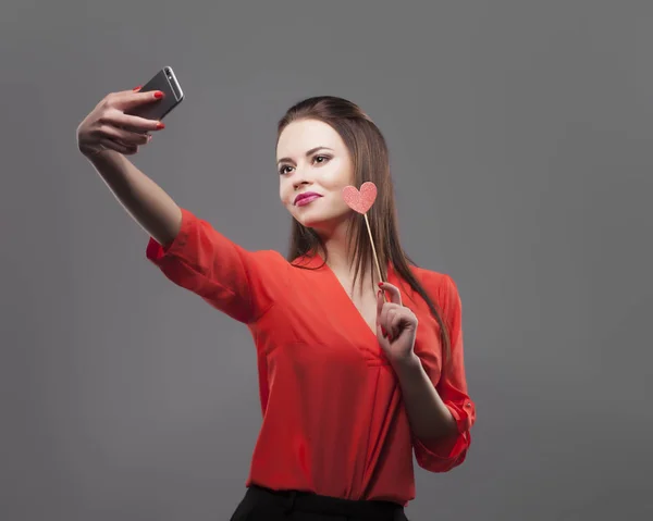 Chica de camisa roja, fondo gris. Joven y alegre morena de moda haciendo selfie con smartphone, sostiene el corazón de papel. Sensación de emoción positiva, sonriendo . —  Fotos de Stock