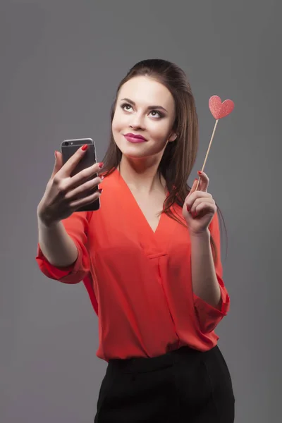 Chica de camisa roja, fondo gris. Joven y alegre morena de moda haciendo selfie con smartphone, sostiene el corazón de papel. Sensación de emoción positiva, sonriendo . —  Fotos de Stock