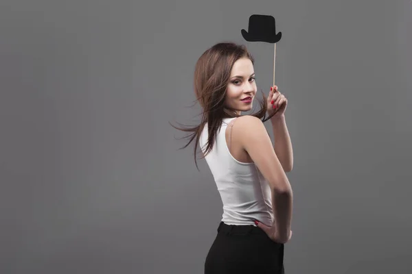 Chica sosteniendo sombrero cilindro de papel divertido en palo, aislado, fondo gris. Joven sonrisa alegre de la mujer de la moda, movimiento del pelo — Foto de Stock