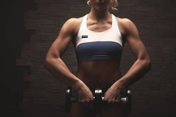 Sexy blond bodybuilder woman in white and blue sports bikini with estonian flag over stone wall background exercising with dumbbells closeup unrecognizable hands shoulders and chest — Stock Photo, Image