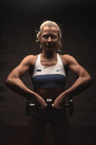 Sexy blond bodybuilder woman in white and blue sports bikini with estonian flag over stone wall background exersising with dumbbells — Stock Photo, Image