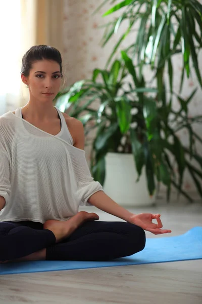 Yoga en casa. Cálmate. Atractiva joven sentada en posición de loto en el suelo con los ojos cerrados en la alfombra azul en camisa blanca —  Fotos de Stock