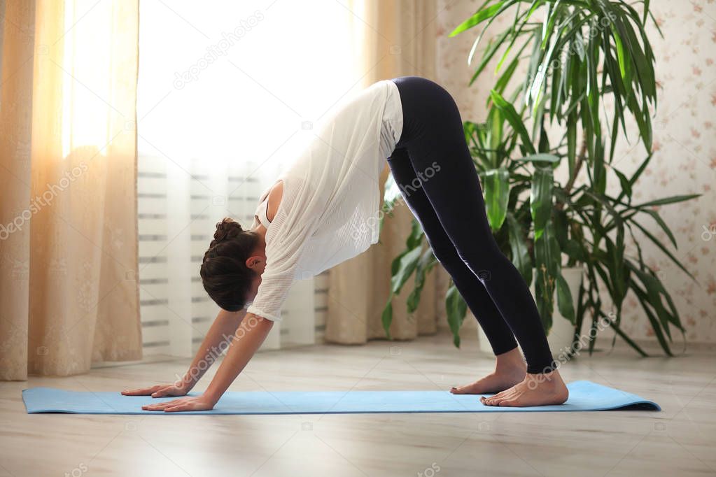 Young attractive woman practicing yoga, standing in adho mukha svanasana exercise, downward facing dog pose, working out, wearing sportswear, white top, pants, indoor full length, home interior