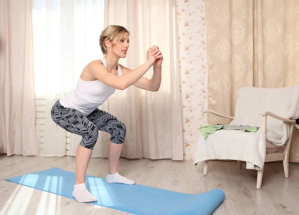 Attraktiv kvinna göra fitness motion hemma på en blå matta i vardagsrummet, gör knäböj — Stockfoto