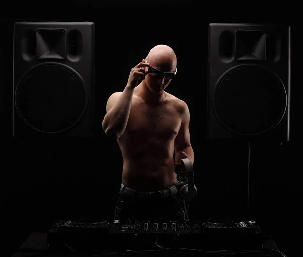 Bold adulto DJ fica perto misturador entre dois grandes alto-falantes seminua com fones de ouvido brancos e óculos de sol — Fotografia de Stock