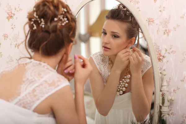 結婚式の準備。室内の白いウェディング ドレスで美しい若い花嫁。鏡を見て、大きな窓付きのスタジオ ルームでイヤリング、自宅で履くはぜモデル. — ストック写真