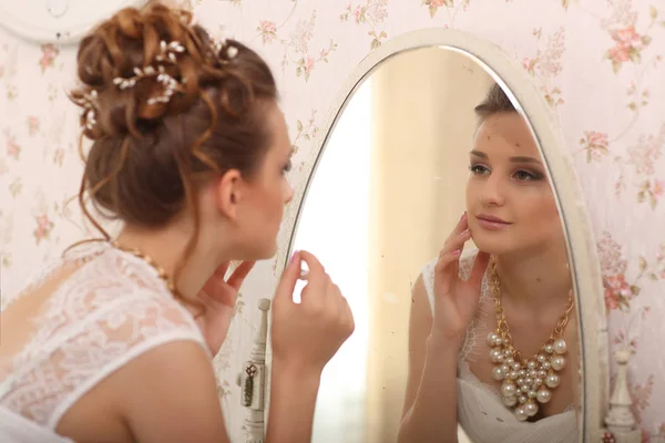 Wedding voorbereiding. bruid in witte trouwjurk binnenshuis. Luxuty model spiegel te kijken en zet op earring, thuis, in de studio kamer met groot raam. — Stockfoto