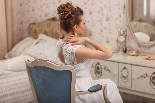Preparazione del matrimonio. Bella giovane sposa in abito da sposa bianco al chiuso. Modello di lusso seduto su una sedia blu vintage a casa — Foto Stock